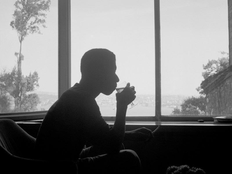 James Baldwin, Istanbul, c. 1965. A reflective moment captured by Sedat Pakay in a gelatin silver print, showing Baldwin’s silhouette seated by a large window, sipping a drink and smoking, with a serene landscape view in the background.(Courtesy of Sedat Pakay)
